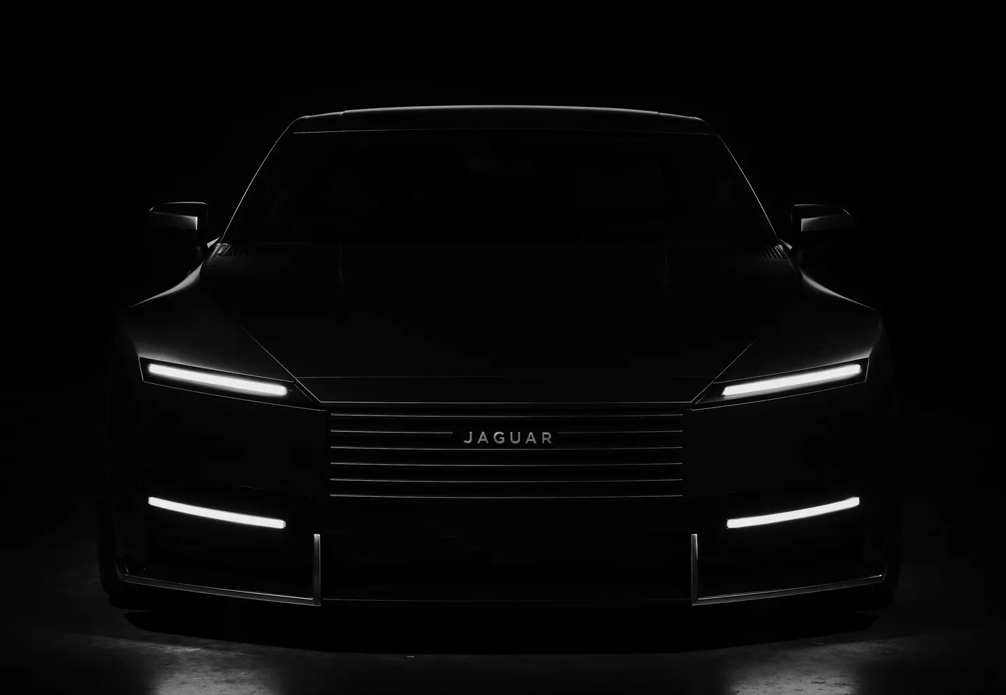 Front view of a sleek black Jaguar Type car in a dark studio with LED headlights and illuminated horizontal grille lines, highlighting its glossy paint and chrome accents.