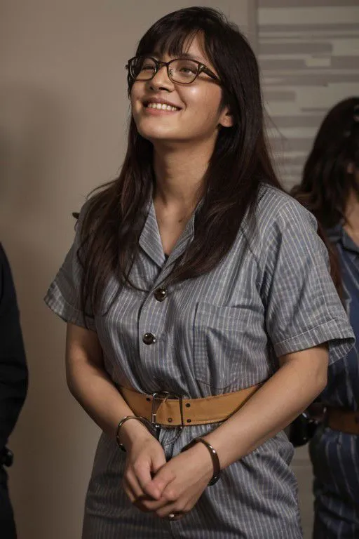 Teen Asian girl wearing rectangle glasses and a striped prison jumpsuit smiling while standing with handcuffs in a courtroom setting