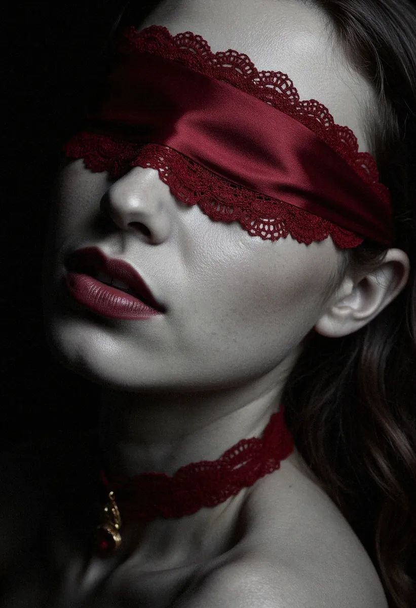Close-up grayscale portrait of a young woman with ivory skin wearing a lace-edged red silk blindfold, maroon lipstick, and a crimson choker with a golden ruby pendant.