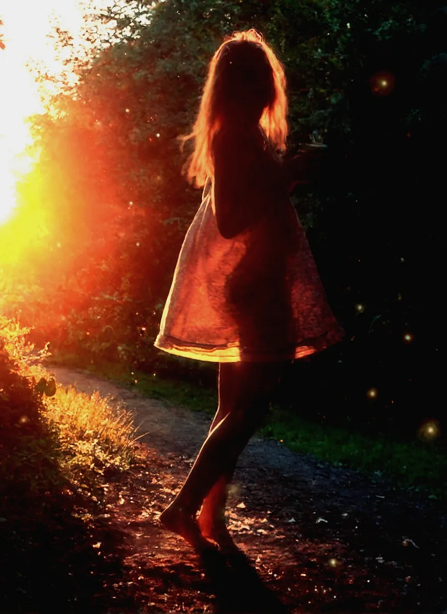 Silhouette of a barefoot girl with long hair wearing a dress, holding a cup and standing outdoors on a path during sunset with strong backlighting and glowing light particles.