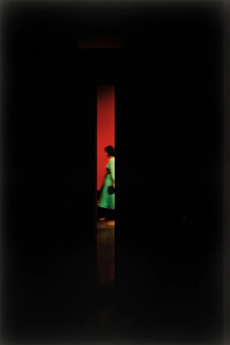 Abstract photograph inspired by Saul Leiter showing a blurry woman in a bright green dress visible through a narrow vertical gap in a dark foreground against a deep red wall.