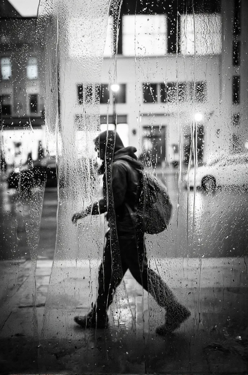Black and white photograph of a person walking away behind a rain-covered frosted glass window with blurred cityscape background