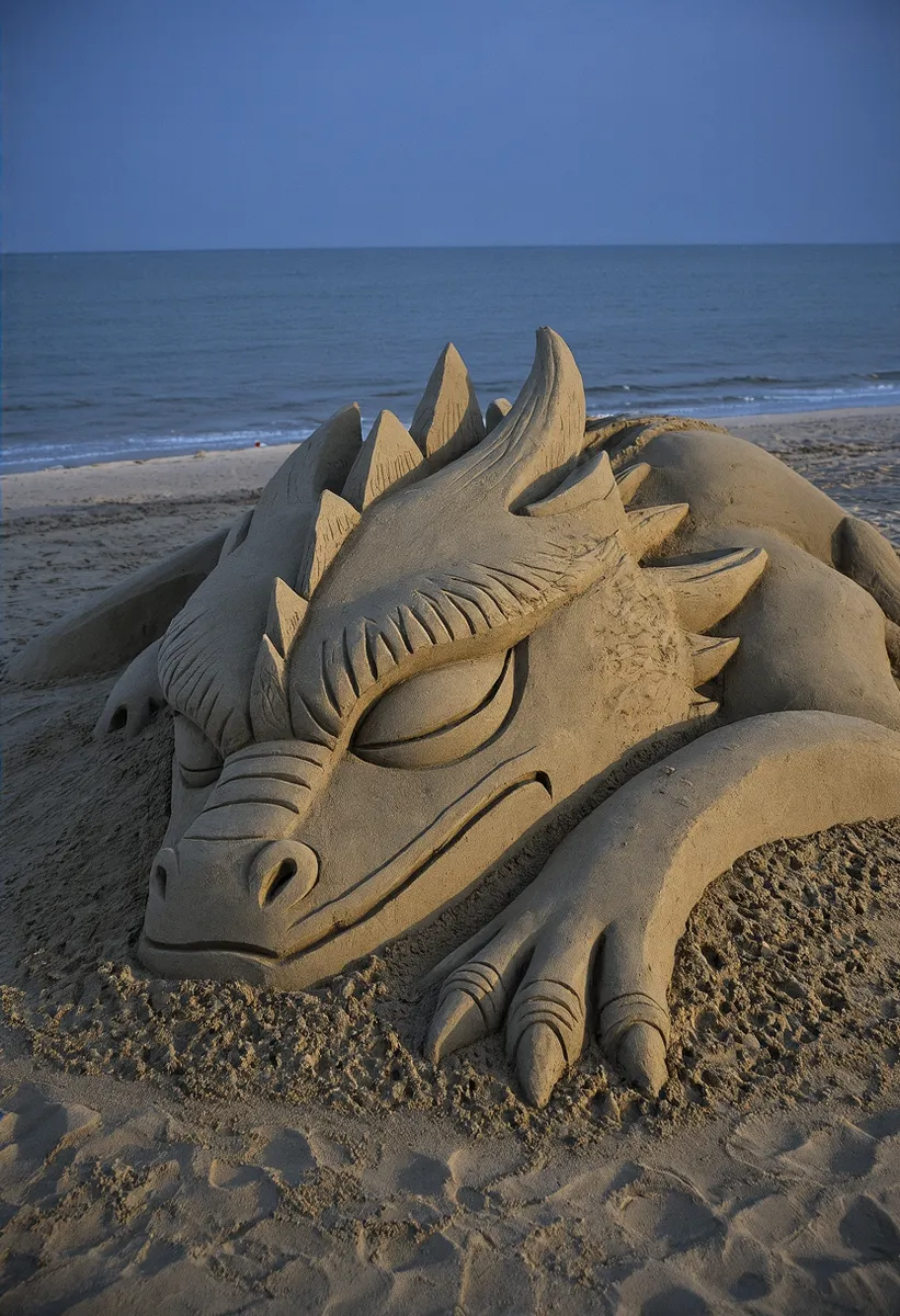 Detailed sand sculpture of a sleeping dragon head on a night beach, showcasing intricate carving with claws and spiked features in temporary art form.