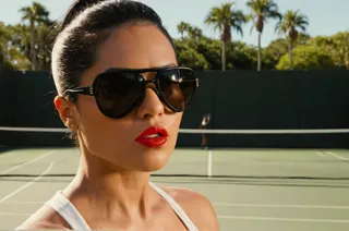 Close-up of a woman wearing black luxury sunglasses and red lipstick, standing on a sunny tennis court with palm trees in the background.