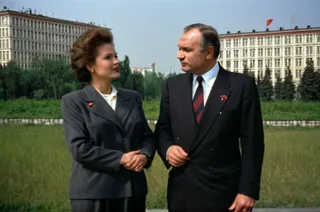 A woman politician and a man stand side by side wearing formal suits with serious expressions, against a backdrop of socialist Soviet housing buildings.