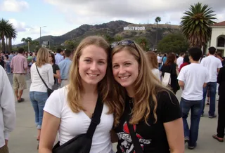 Two blonde women posing and smiling in front of the Hollywood sign in Los Angeles on a busy street with tourists around.