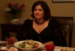 Woman in her 40s wearing a lavish black dress and necklace, sitting at a dining table with a salad bowl and red drink, warm indoor lighting