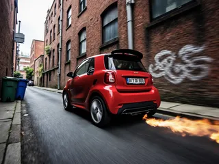 Red smart car with a spoiler driving fast in a narrow back alley with brick walls and graffiti, producing flames from its exhaust pipe with motion blur effect.