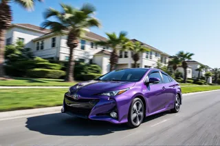야자수와 대저택이 있는 주거 거리에서 빠르게 달리는 보라색 Toyota Prius, 배경에 맑은 파란 하늘과 모션 블러 효과.