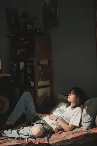 Photorealistic image of a young woman lying on a bed indoors, bathed in soft, natural light highlighting her detailed face and casual clothing.