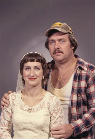 A vintage bridal portrait from 1979 featuring a bride in a lace wedding dress with heavy makeup, and a middle-aged groom in a trucker cap and unbuttoned flannel shirt.