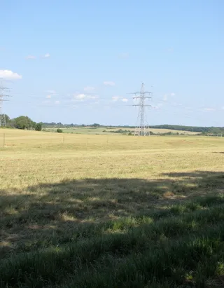 A simple rural field with green and yellow grass under a clear blue sky, featuring metal transmission line pylons in the distance.