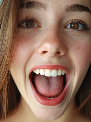 Close-up photograph of a young woman with brown eyes and long hair, mouth wide open showcasing perfect symmetric teeth and detailed tongue texture.