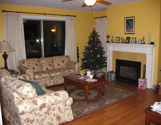 Cozy 2000s living room with floral patterned sofas, a decorated Christmas tree in the corner, a fireplace with decorative items, yellow wallpaper, and a wooden coffee table on a patterned rug at night.