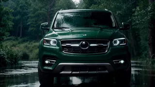 Front view of a luxury emerald green SUV with illuminated aggressive headlights and large front grill, parked on wet ground with raindrops, forest background under cloudy sky.