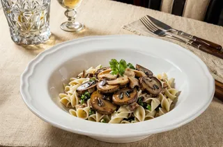 A ceramic bowl filled with Farfalle pasta topped with sautéed mushrooms and garnish, placed on a beige cloth with a glass, cutlery, and a vase holding a red rose in dim evening lighting.