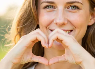 Close-up soft-focus portrait of a young woman smiling gently while forming a heart shape with her hands at chest level, with warm lighting and a blurred bokeh background.