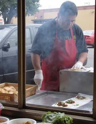 Mexican American man in red apron serving tacos at a taco stand on a Los Angeles street, sidewalk and parked cars visible, steam rising from grill