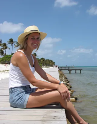 Woman in her 20s with blonde hair wearing a straw hat, white tank top, and jean shorts sitting on a wooden pier by the ocean on a sunny day in Florida.