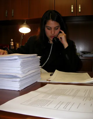 A lawyer in a black suit and black hair seated at a wooden desk in an office, holding a phone, surrounded by large stacks of paperwork and documents under warm LED lighting.