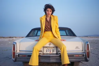 Woman in yellow blazer and bell bottoms sitting on the trunk of a white Cadillac car with Utah license plate at blue hour in salt flats, windy weather.