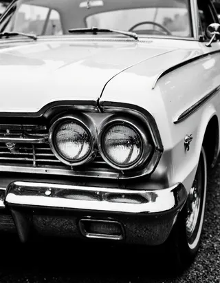 Black and white close-up photo of a vintage 1960s car front showcasing dual round headlights and chrome bumper.