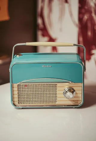 A retro style portable blue radio with a beige handle and a large knob tuner sits on a white surface indoors.