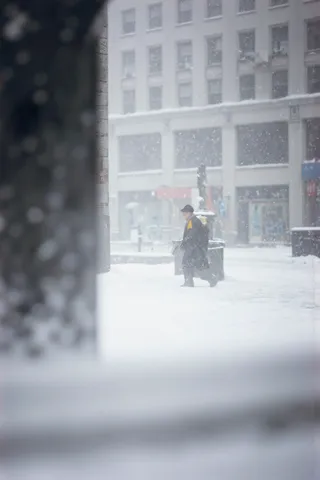 A solitary man walks on a snow-covered urban street during heavy snowfall, partly obscured by a large dark blurred foreground object, with a bright yellow scarf detail amid soft gray and white cityscape.