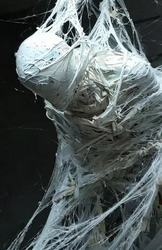 A body tightly wrapped in white spider web-like fabric, suspended with intricate weaving against a dark background, illuminated by soft light highlighting texture.
