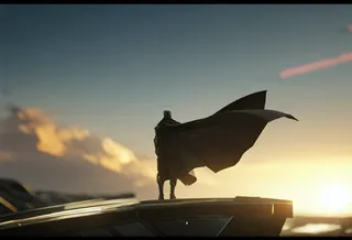 Faceless male figure wearing a futuristic suit and a flowing long cape standing on a platform at sunrise with a huge alien spaceship in the blurry background, viewed from behind.